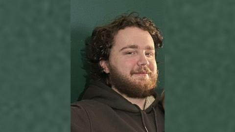 Man with curly brown hair and brown beard, smiling and looking at the camera with a dark green wall in the background