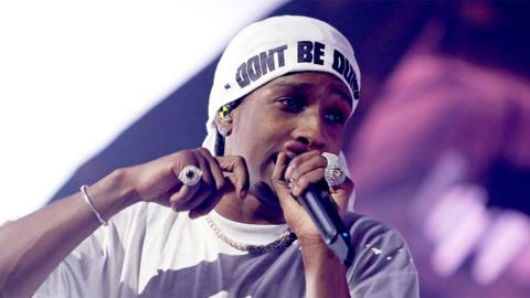 A$AP Rocky performing on stage, singing into a black microphone while wearing a white tshirt, white hat which reads "Don't be Dumb", silver chain around his neck  and sparkling rings on his fingers.