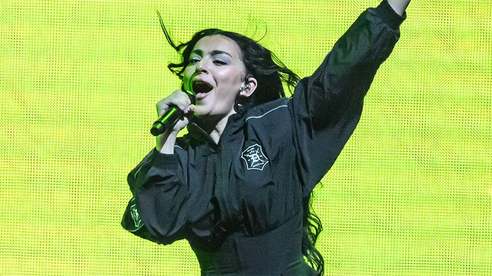 Charli XCX, a woman, on stage with a microphone in her right hand, wearing black outfit with the background screen a lime green colour.