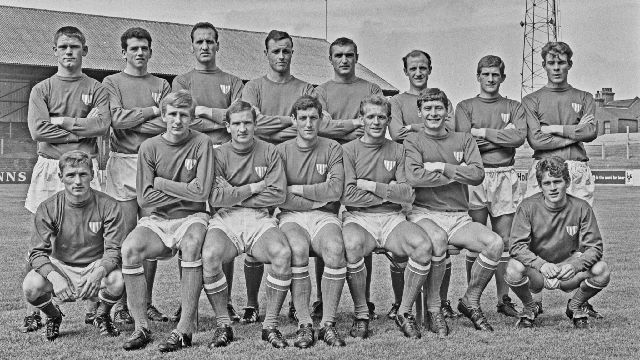 A picture of the Leyton Orient League Division 2 team in 1965 in their full football kit. There are two rows of players in white shorts and long sleeved jerseys. The goalkeepers are crouched at either end. Colin Flatt is in the middle of the picture on the front row.