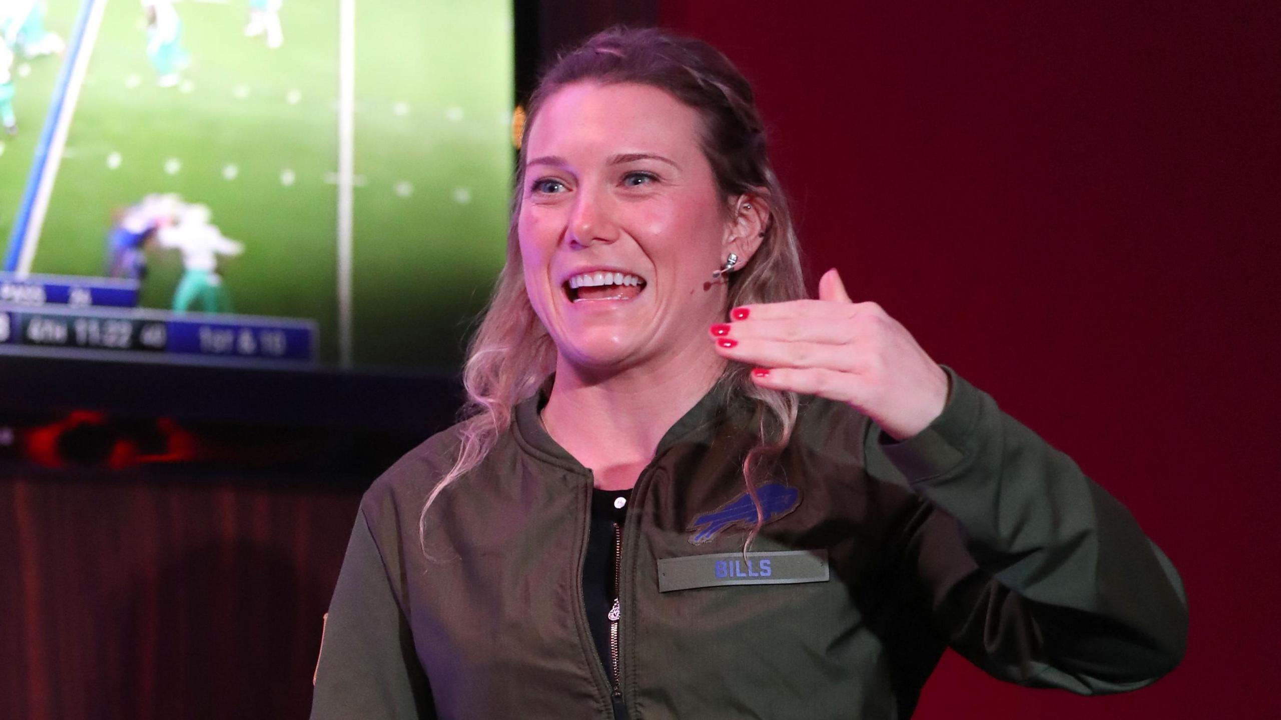 Phoebe Schecter wearing a Buffalo Bills jacket with a screen showing NFL action behind her