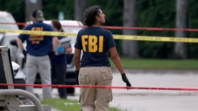 An FBI agent with the letters 'FBI' on the back of their T shirt looks on as they walk between police tape with  two other officials in the background, in West Palm Beach, Florida on 15 September