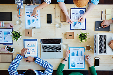 An overhead camera shot of a group of people sitting at a table. The table is a natural piece of wood. All but one person has a clipboard and a printout in front of them containing various bar charts and pie charts. One individual, sitting at the head of the table, has a laptop in front of them. There are also various mobile phones, pot plants and mugs of drink on the table.