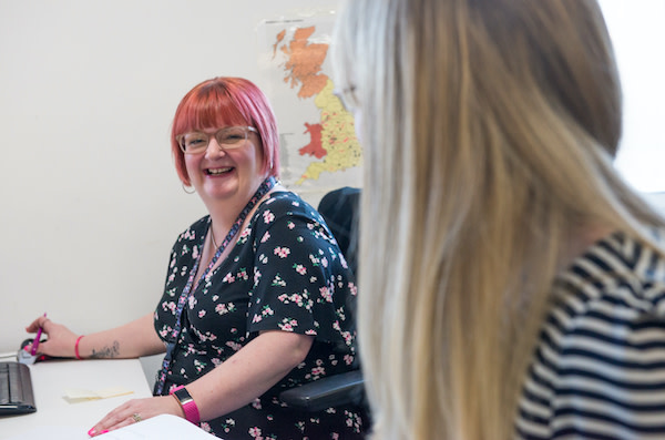 Two employees sit side by side at their desks within an office. One employee is turned sideways to the camera, where their face cannot be seen, the other is looking at the camera and smiling.