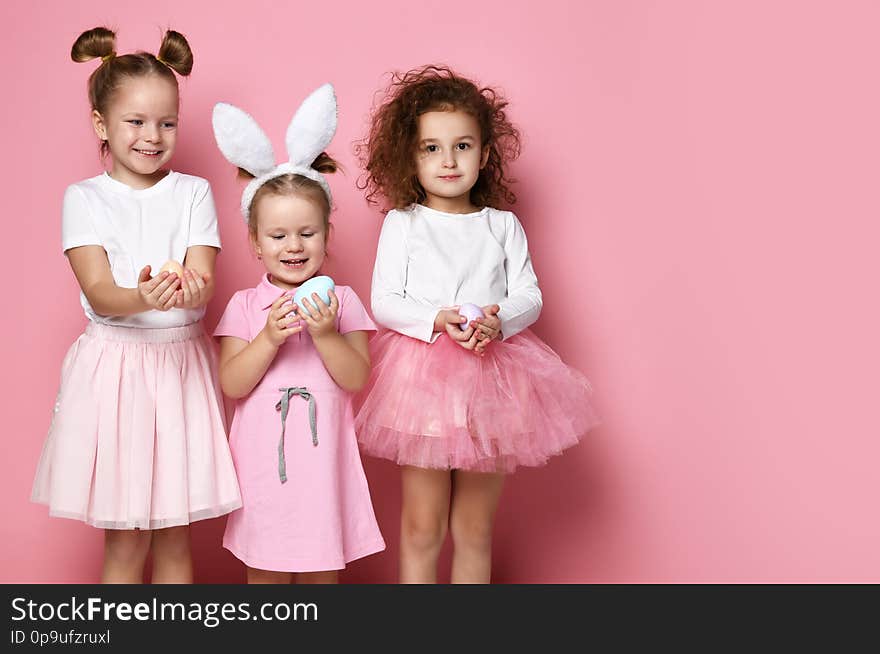 Three smiling dressed up for the holiday kid girls hold painted eggs on Easter day. Happy Easter celebration on pink background with free text space