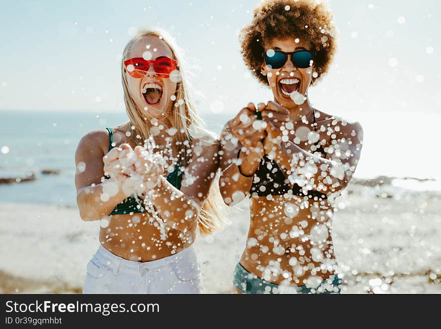 Excited young women friends bursting a water balloon and smiling by the sea. Best friends having fun on beach holiday