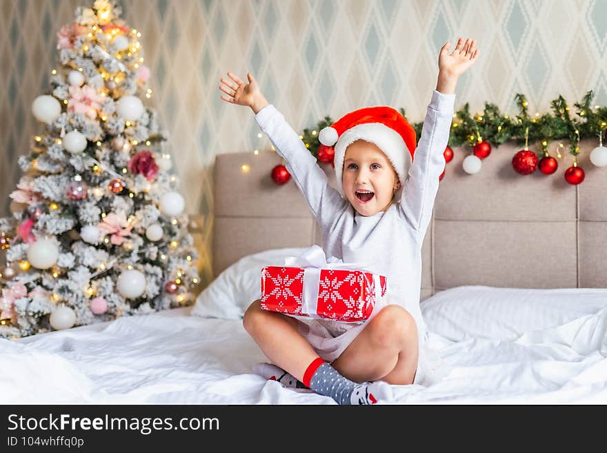 Preschool funny girl sit on bed near decorated Christmas tree in beautiful room in the holiday morning, enjoing with presents.