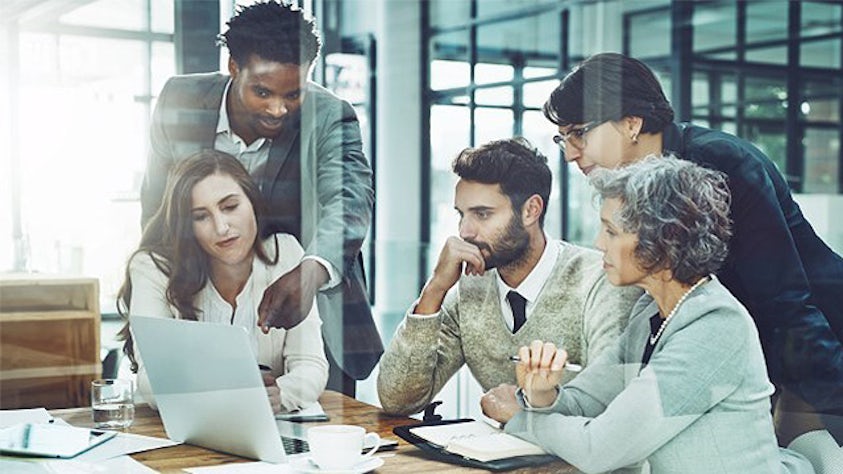 Group of people in an office gathering around a laptop on a conference table