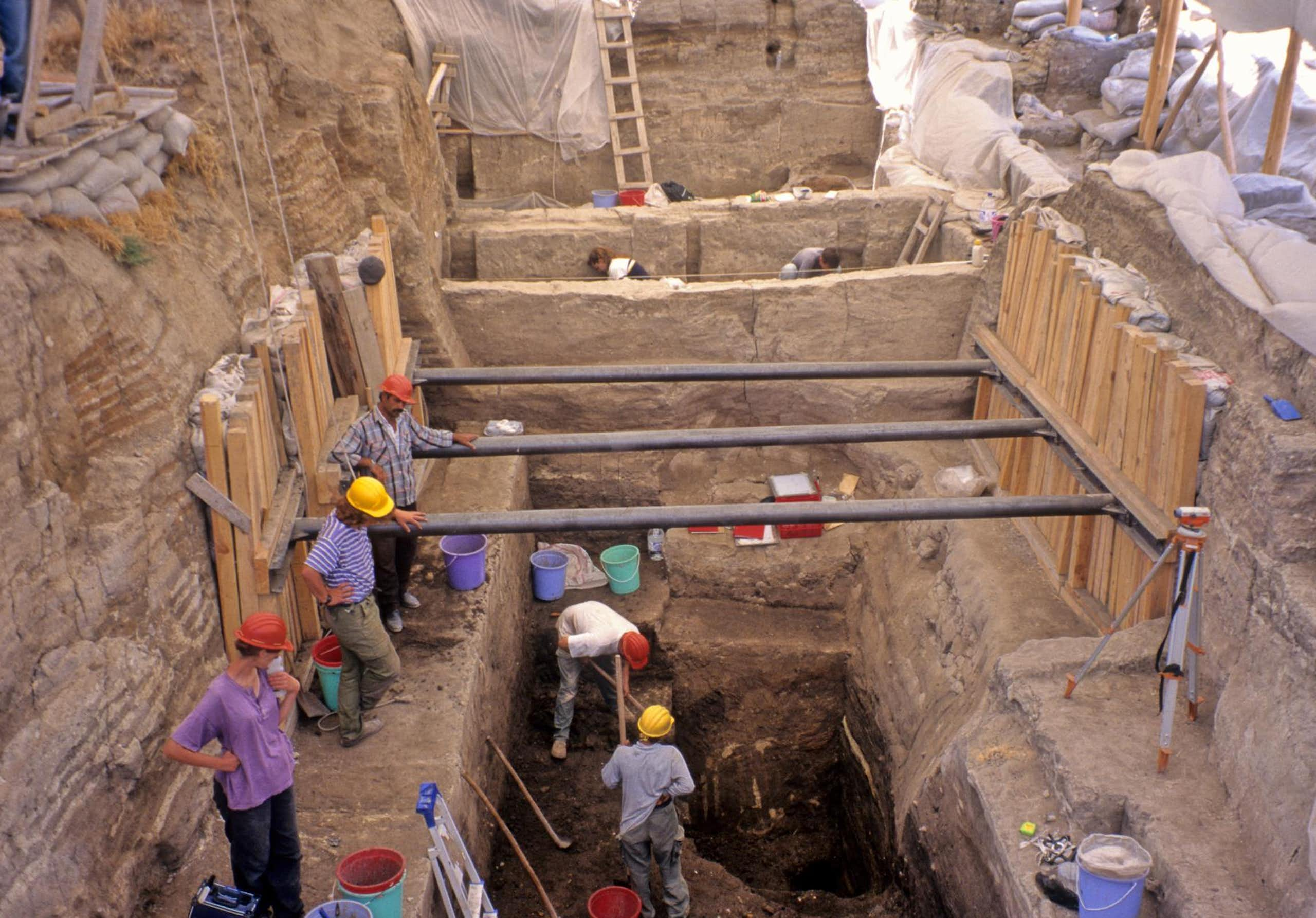 people in hardhats stand along perimeter of a trench in dusty land, a couple dig within