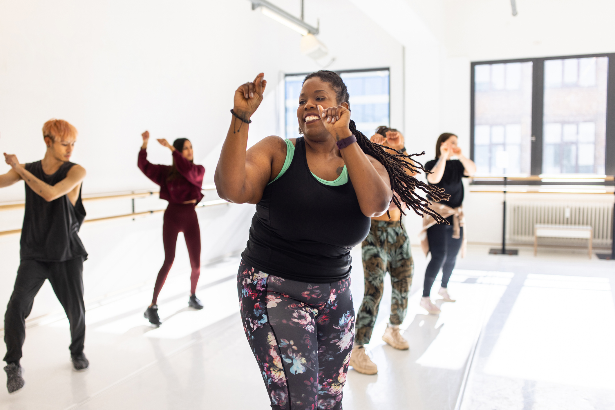 People dancing energetically in a bright studio, led by a person with long braided hair wearing a black tank top and floral leggings