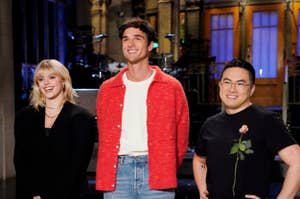 Chloe Fineman, Michael Longfellow, and Bowen Yang stand together smiling; Fineman in a black blazer, Longfellow in a red cardigan, and Yang in a black shirt with a rose print