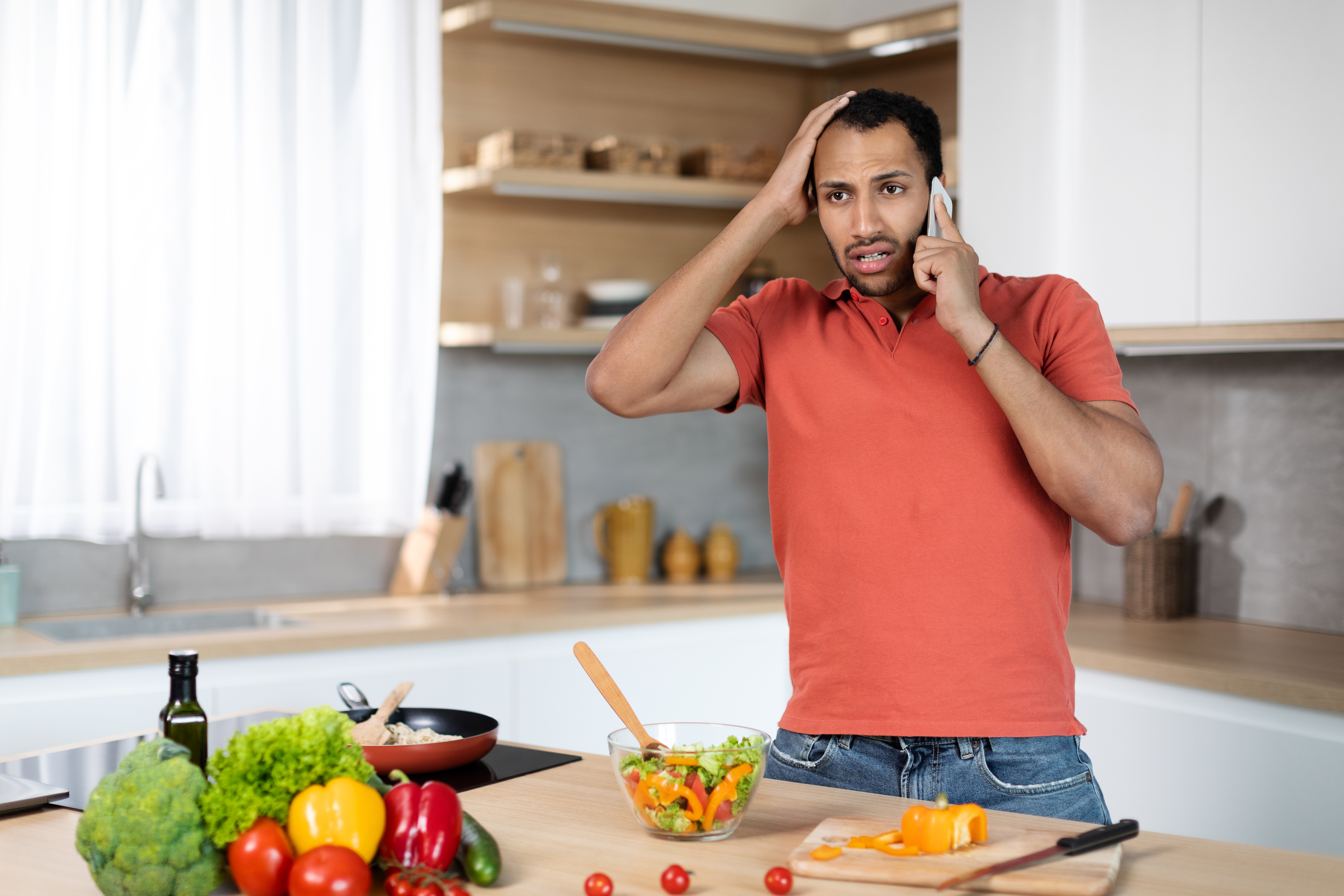 A man in a kitchen looks stressed while talking on the phone, surrounded by fresh vegetables and a salad in progress