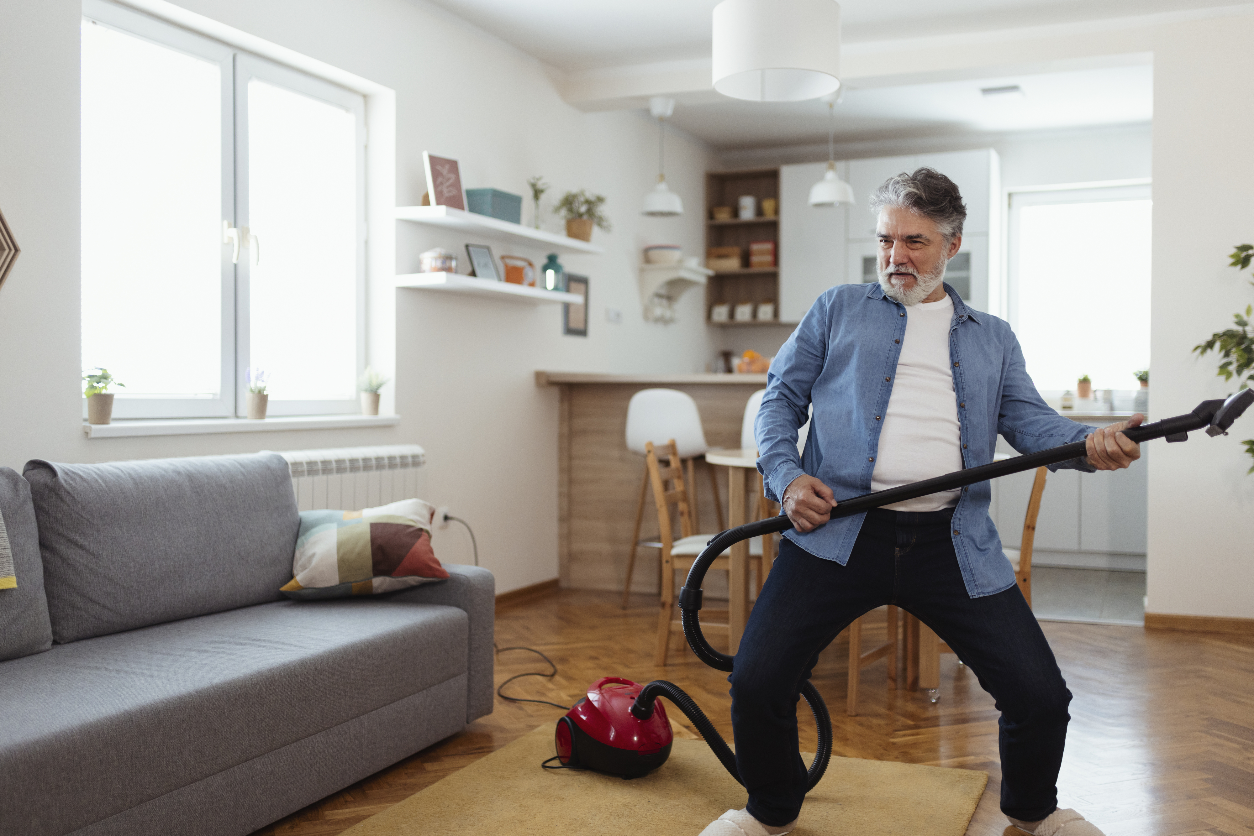 A man playfully holds a vacuum cleaner like a guitar while standing in a cozy, modern living room. He is smiling and appears to be enjoying himself