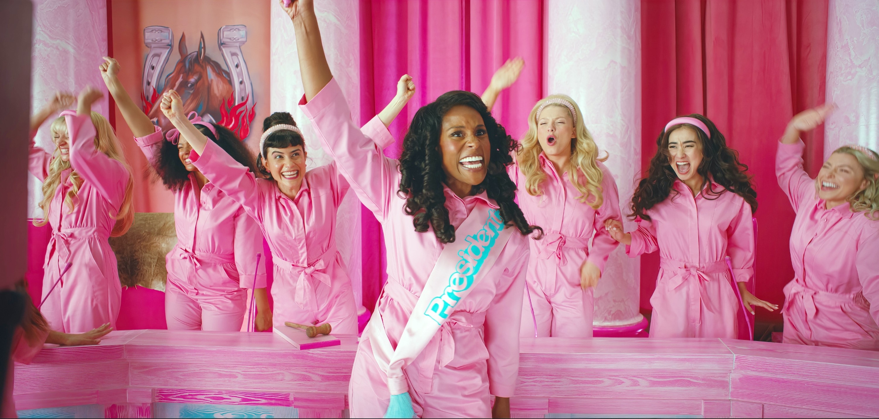 Women in matching pink outfits, including Issa Rae in a &quot;President&quot; sash, cheer together in a lively scene with a pink background