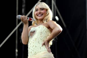 A woman with long blonde hair in a sparkly strapless dress holds a decorated microphone and smiles at an outdoor event