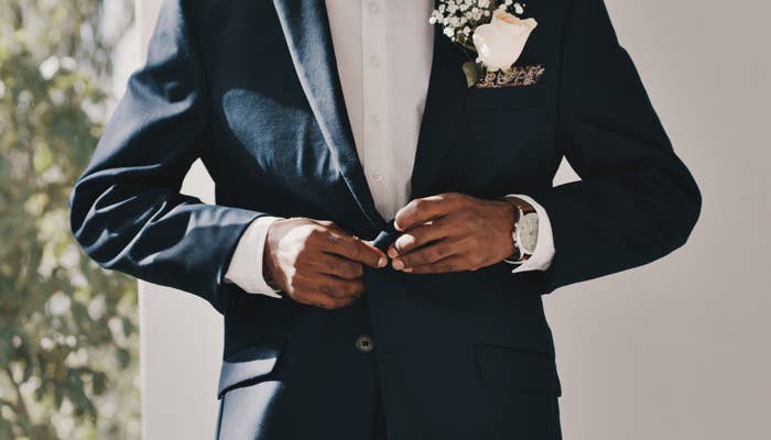 A groom in a suit adjusts their jacket, showcasing a boutonniere with flowers and a watch on their wrist