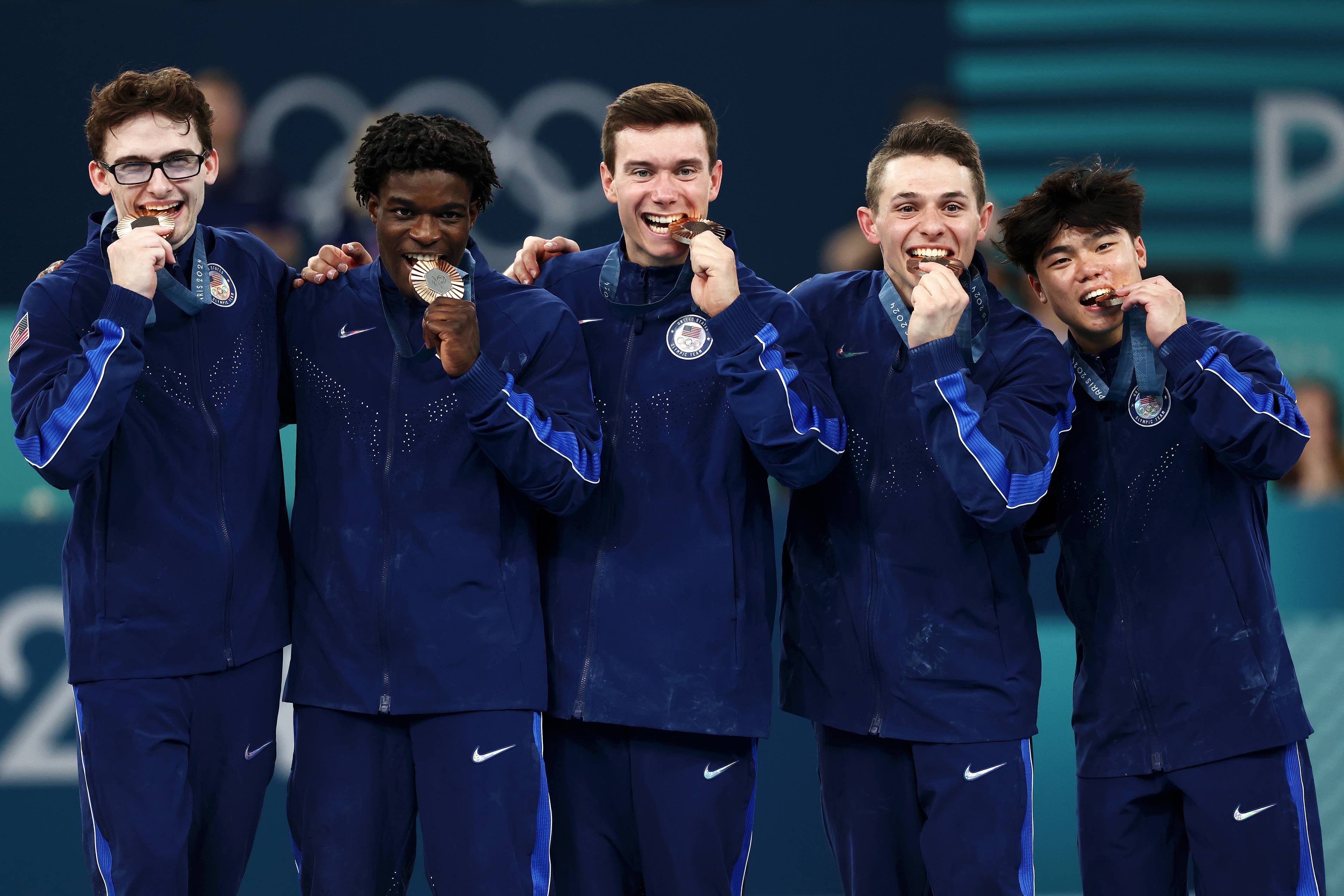 Group of five male athletes in tracksuits, each biting a medal and standing together, celebrating