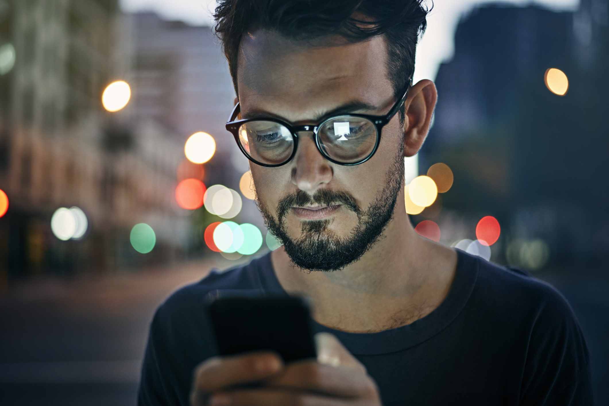 Man with glasses looking at his phone on a city street at night; background shows blurred lights