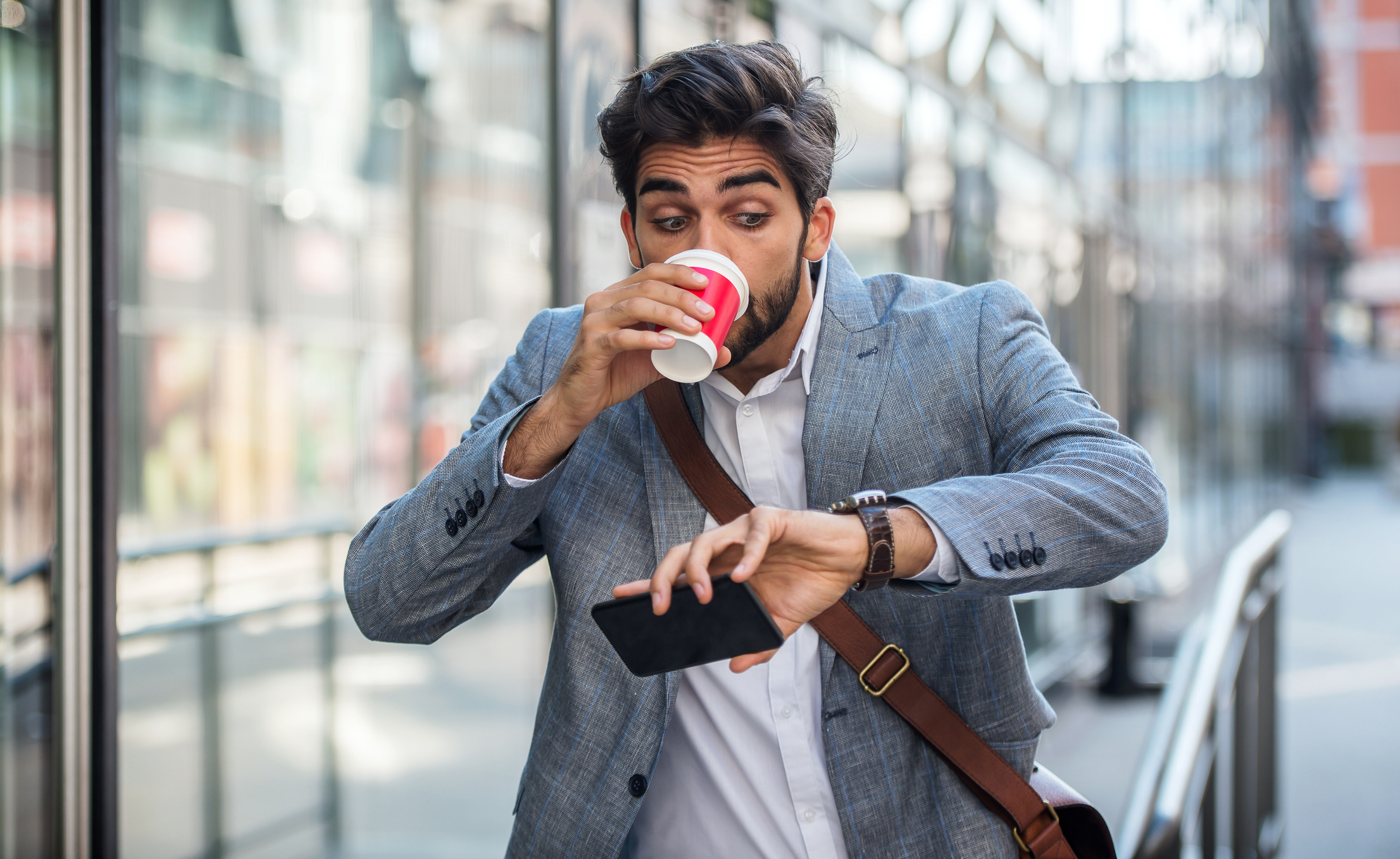 Man in a blazer drinks from a cup while checking his watch and holding a phone in a busy, outdoor urban area