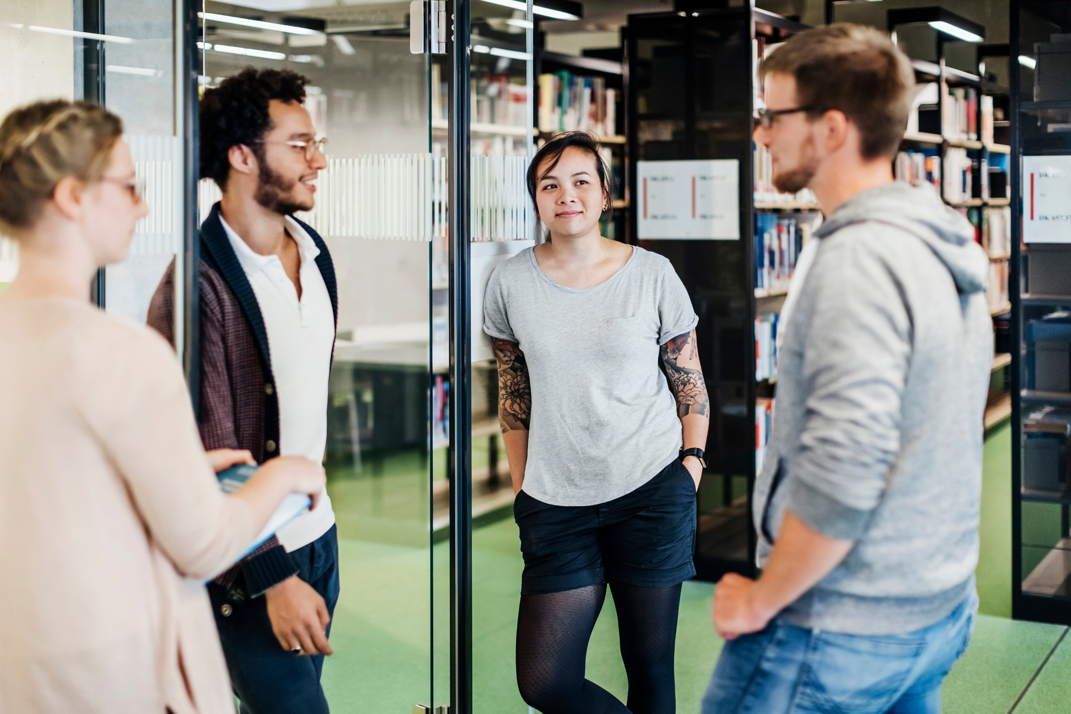 Four people stand and chat casually in a library. All are dressed in casual clothing