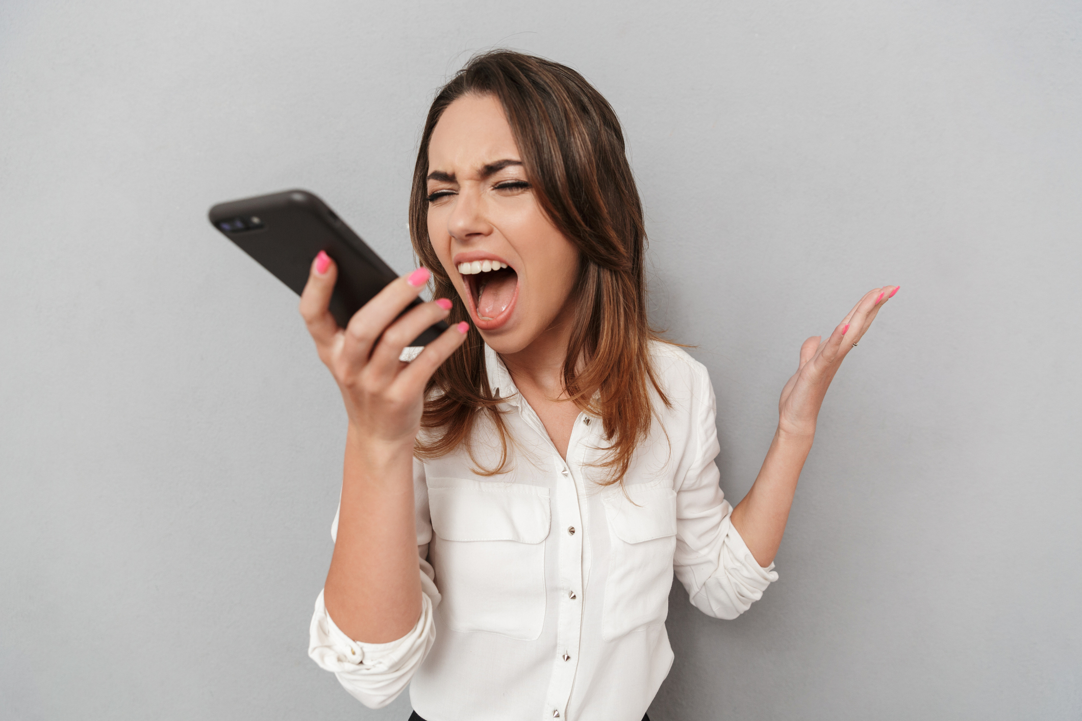 A woman in a white blouse is yelling into her smartphone with an angry expression while gesturing with her other hand