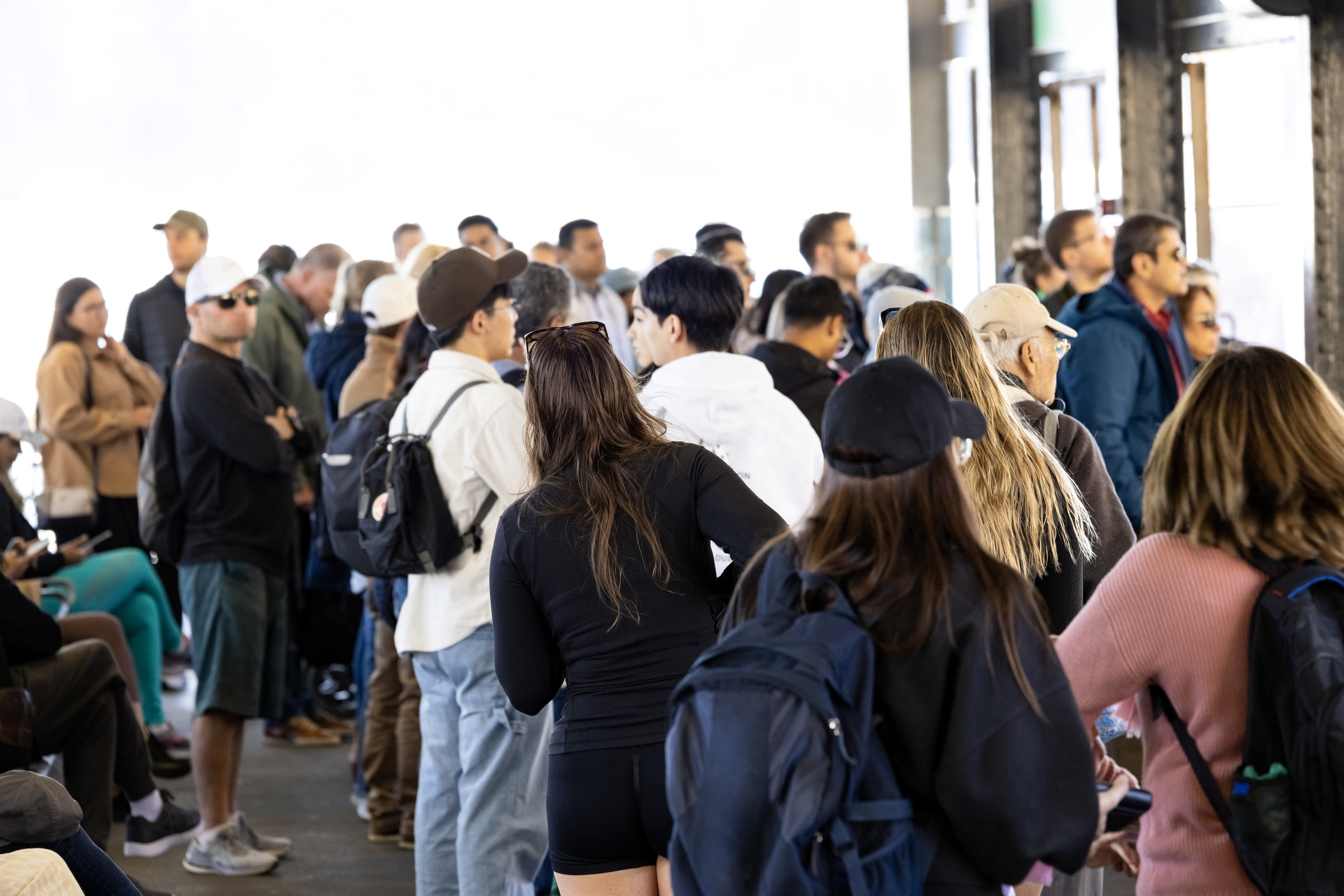 A crowded scene with many people in casual clothing standing and waiting, some with backpacks and hats. The setting appears to be a waiting area