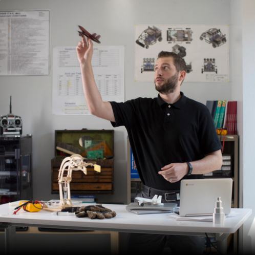 A male teacher stands behind his classroom desk, holding a model airplane in the air with one hand.