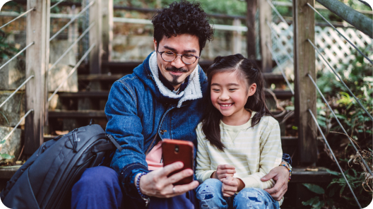 A father and daughter look at a phone together, smiling. They are sitting outside on steps.