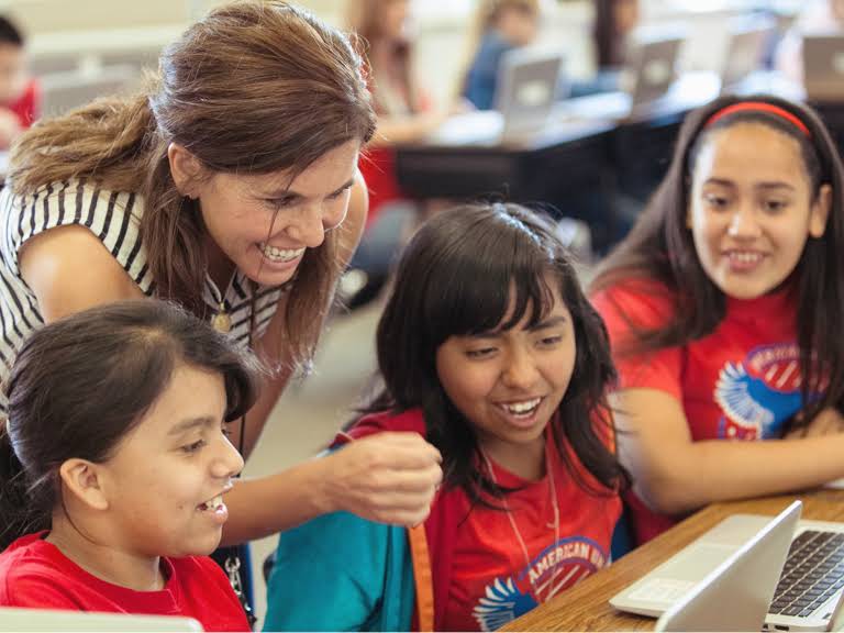 A smiling teacher looking at something on a screen with three seated female students.