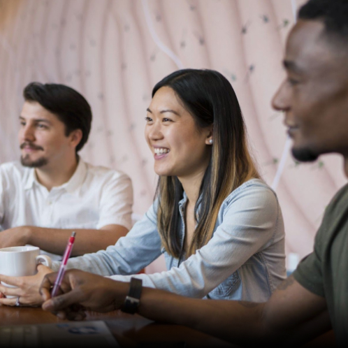 Three young adult students sit at a long desk, smiling and listening to someone out of the frame speak.