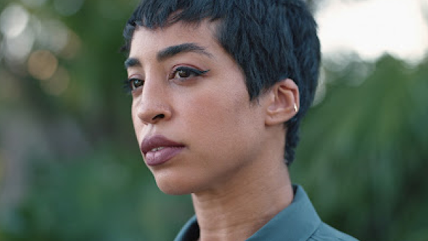 Jillian Mercado (she/her), a Black disabled woman, wears a teal shirt and looks into the distance