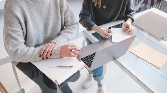 Two people work while sitting next to each other at a glass desk with a laptop.