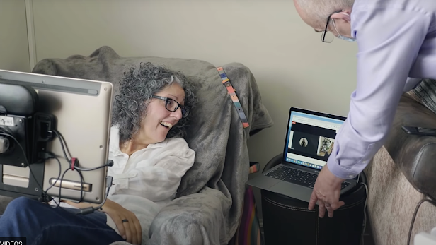 A woman with grey hair and black glasses sits in a chair with a screen in front of her looks to the side smiling