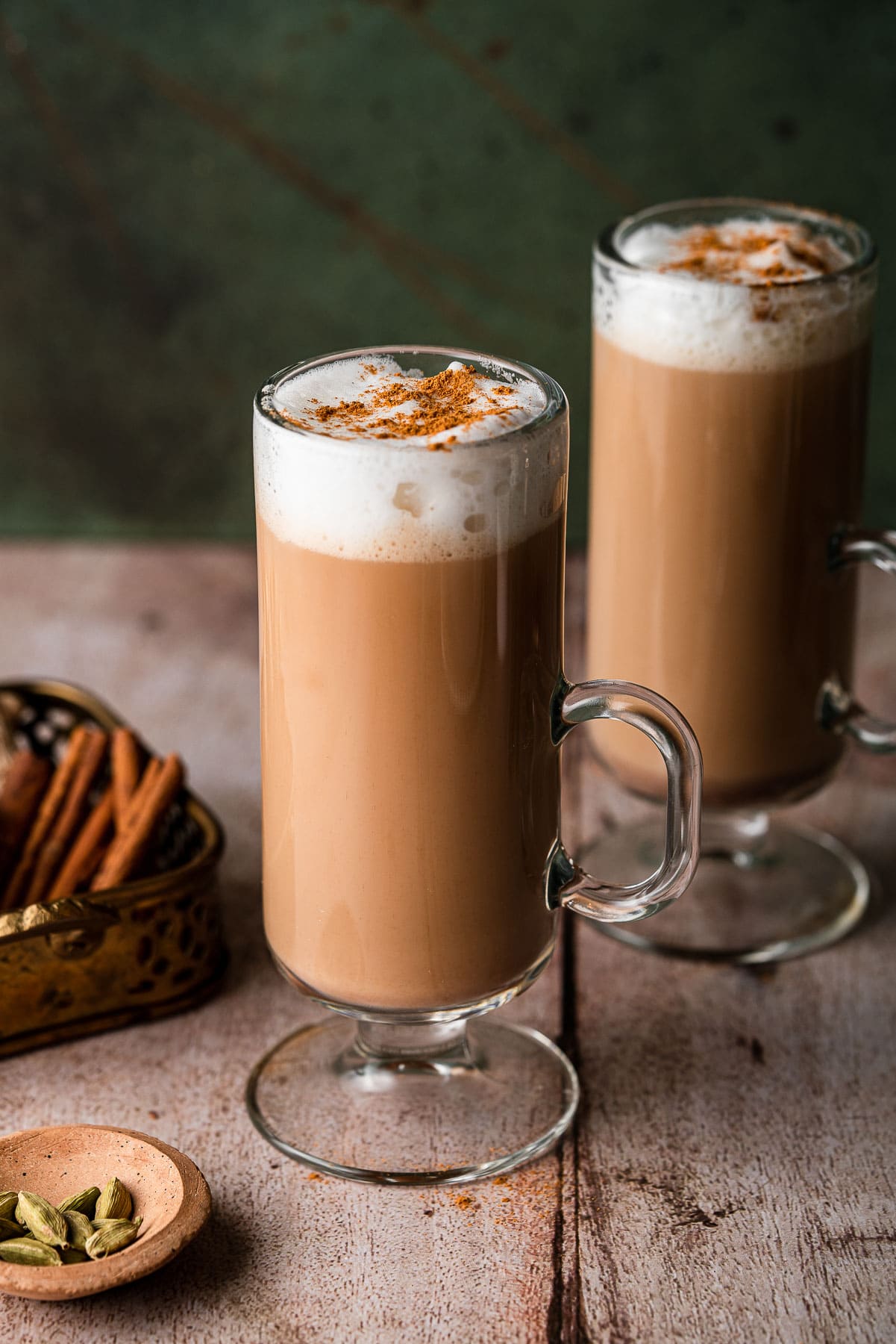 two irish coffee mugs with foamy chai tea lattes with whole cinnamon sticks and cardamom pods.