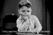 a black and white photo of a little boy sitting at a table with his hand on his chin .