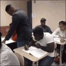 a group of people are sitting at desks in a classroom while a man stands in front of them .