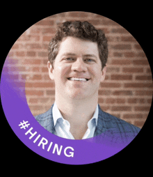 a man is smiling in front of a brick wall and a purple circle with #hiring written on it