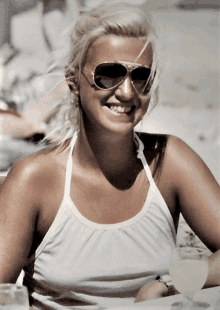 a blonde woman wearing sunglasses and a white tank top smiles