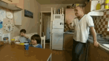 a man leans on a counter in a kitchen while two children sit at a table with a blue cup on it