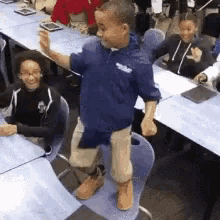 a boy in a blue shirt is standing on a chair in front of a group of children sitting at tables .