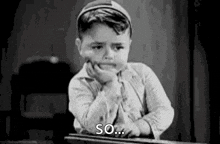 a black and white photo of a young boy sitting at a desk with his hand on his face .