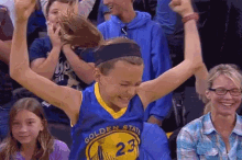 a girl wearing a golden state warriors jersey is sitting in the stands with her arms in the air .