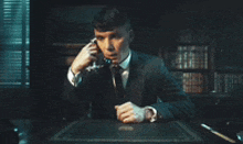 a man in a suit is sitting at a desk talking on a cell phone .