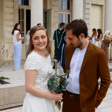 a woman in a white dress stands next to a man in a brown suit holding a bouquet of flowers