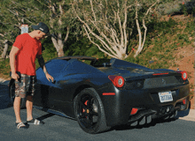 a man in a red shirt stands next to a black sports car with a california license plate that says tx7z064