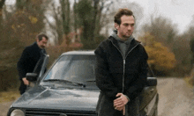 a man in a black jacket is standing next to a car on a dirt road .