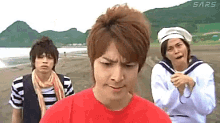 a group of young men are standing on a sandy beach and one of them is wearing a red shirt