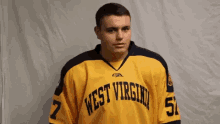 a man wearing a yellow and black west virginia hockey jersey is standing in front of a white wall .