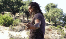 a man with long hair and a tattoo on his arm is standing in a field with trees in the background