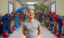 a man wearing glasses is standing in front of a painting of superheros standing in a hallway .